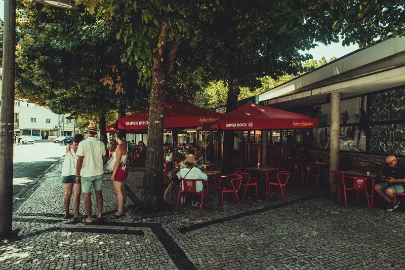 Outdoor café with red umbrellas, people relaxing under trees. Summer day in urban setting.